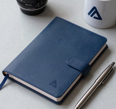 A high-quality assortment of corporate gifts including a dark blue agenda, a metal pen, and a ceramic mug, all featuring a professional logo, set against a light grey surface.