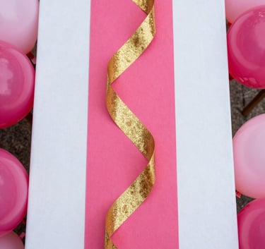 Close-up overhead flat-lay of a styled party tablescape — hot pink and gold layered accents, sparkle ribbon, balloon clusters at frame edges, bright studio lighting, saturated color, no people