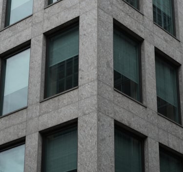 A close-up, minimalist architectural photograph of a sleek office building corner in North America, focusing on textures of gray stone and dark glass with soft teal reflections.
