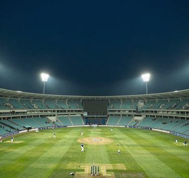 A panoramic photography of a massive cricket stadium in India during a night match, illuminated by intense teal and white lights against a deep dark blue sky, viewed from the stands.