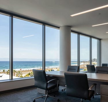 A wide-angle professional photograph of a luxury North American / US office interior in Huntington Beach, featuring large windows overlooking the Pacific coast, decorated in steel blue and soft grey tones.