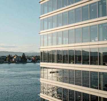 A panoramic photograph of a modern glass office building in Kirkland, Washington, reflecting a clear light blue sky and the calm waters of Lake Washington. The composition is wide and professional, conveying a sense of stability and North American corporate excellence. Palette tones are predominantly muted blue and off-white.