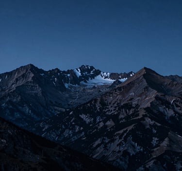 A wide-angle shot of the Pyrenees mountain range at dawn. The peaks are silhouetted against a deep midnight blue sky with hints of soft steel blue light on the ridges. Atmospheric and serene.