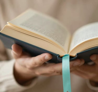 A close-up, elegant shot of a person's hands holding a book manuscript, with a soft turquoise ribbon bookmark. The lighting is warm and inspiring, suggesting a sacred work. Colors: #A1D4D0, #C7A196, #FDF7F5.