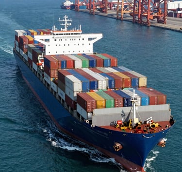 A high-angle professional photograph of a massive container ship loaded with colorful shipping containers navigating a deep blue ocean toward a modern international port. Bright daylight, crisp details, representing global logistics and reliability. Global / International context.