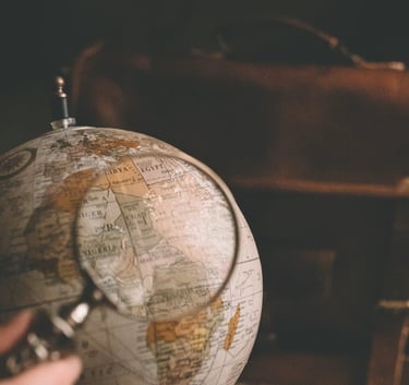 A magnifying glass held over a vintage world globe next to a leather travel bag for adventure planning.