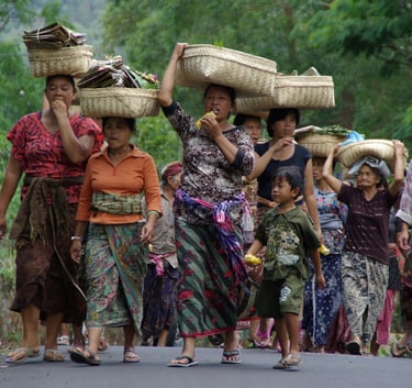 Balinese women in traditional sarongs carrying woven baskets on their heads while walking on a rural road.