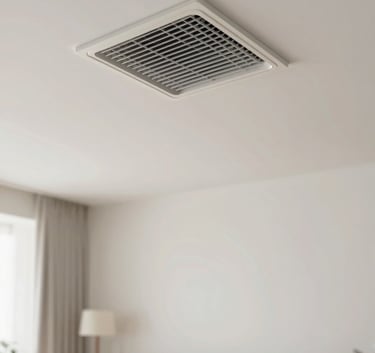 An indoor air conditioning vent on a ceiling in a contemporary North American / US living room. The room below is blurred but shows soft off-white furniture, suggesting a cool and relaxing atmosphere. The lighting is natural and clear.