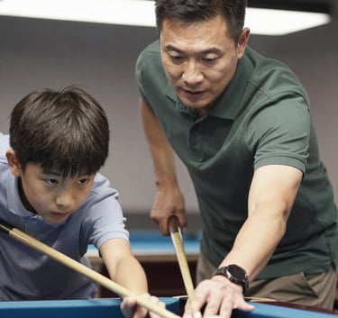 A professional mentor in a dark slate green polo shirt teaching a young student how to line up a shot on a billiard table. The lighting is bright and inviting, showcasing a positive learning environment.