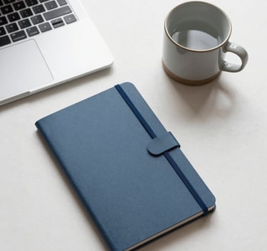 A top-down view of a minimalist desk with a laptop, a Slate Blue notebook, and a ceramic mug on a Snowy Off-White surface.