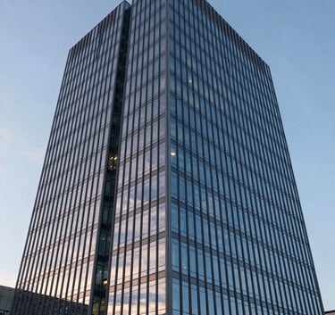 A wide-angle shot of a modern glass corporate skyscraper reflecting a soft sky blue sky at dusk. The architecture is sharp and clean, with muted steel blue structural elements visible behind the glass.