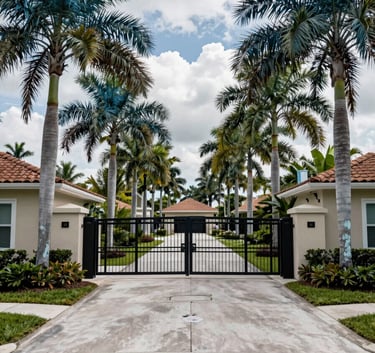 A high-quality photograph of a gated residential community entrance in Florida. Mature palm trees in deep teal pine tones line the drive. The image conveys a sense of safety and professional management.