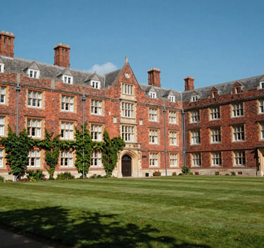 A beautiful wide photography shot of a historic red-brick UK university campus with green ivy and well-maintained lawns under a clear blue sky.