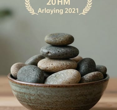 A close-up photograph of a ceramic bowl filled with smooth river stones on a wooden surface, suggesting balance and peace. The background is a blurred soft sage wall.