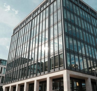 A professional wide shot of a modern glass-fronted office building in a British city, reflecting a clear light blue sky. The architecture is sleek with steel and off-white stone accents, conveying a corporate and trustworthy atmosphere.