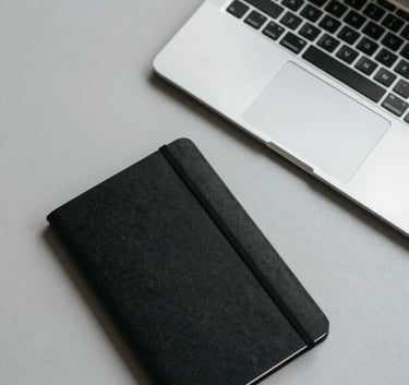 A top-down view of a minimalist desk setup with a sleek laptop and a single black notebook on a light gray surface, professional North American office setting, clean lines and clutter-free.