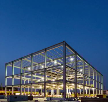 A wide-angle technical photograph of a large-scale commercial construction site at dusk. The structural steel frame is highlighted by warm site lights against a dark royal blue sky. Sharp focus and professional architectural photography style.
