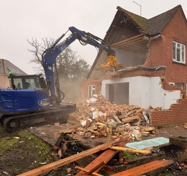 A blue hydraulic excavator demolishes a brick residential house during a site clearance and renovation project.