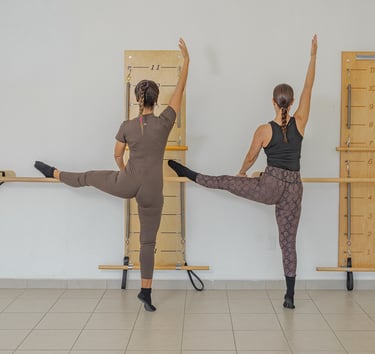 Two women performing a side leg raise stretch against the barre in a Pilates class.