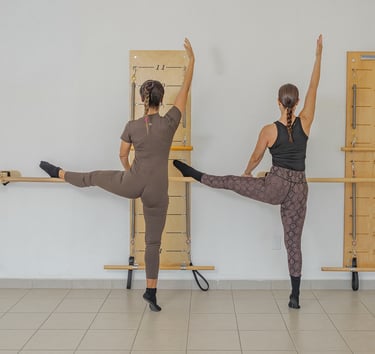 Two women performing a side leg raise stretch against the barre in a Pilates class.