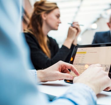 Person using a tablet during a meeting, with colleagues in discussion in the background.