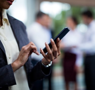 Person using a smartphone with colleagues conversing in the background.