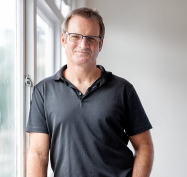 Photo of a man wearing a glasses, a black polo shirt and jeans, leading on a window sill and smiling