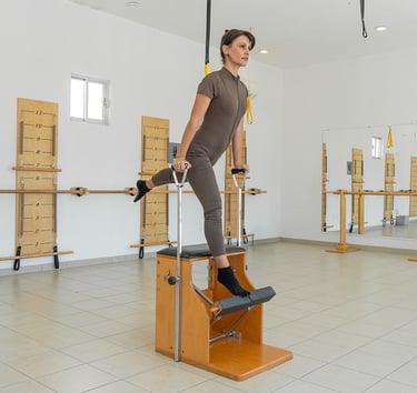 Woman performing a standing leg exercise on a wooden Pilates Wunda Chair in a studio setting.