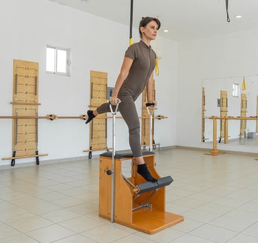 Woman performing a standing leg exercise on a wooden Pilates Wunda Chair in a studio setting.