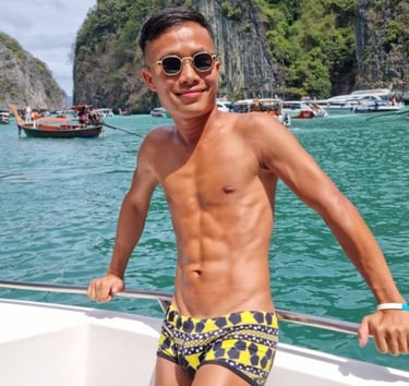 A male outcall masseur in pattaya standing on a boat wearing swim wear with the sea and an island in the background