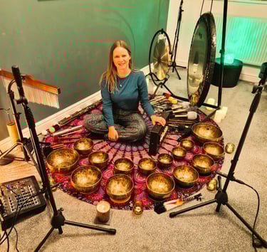 Chrissy sitting on a mat with shruti box, chimes, gongs, many singing bowls, and audio equipment