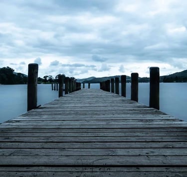 A long wooden pier stretching into a calm blue lake under a cloudy sky with mountain silhouettes.