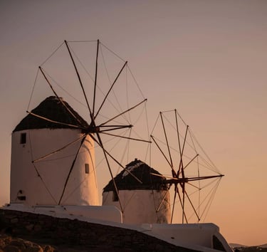 Traditional white Mykonos windmills at sunset, a scenic landmark in Greece with a golden sky.