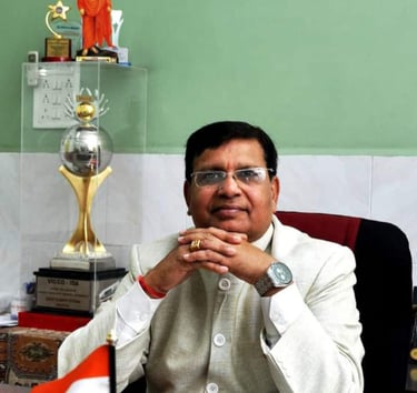 Professional man in white Nehru jacket sitting at a desk with an Indian flag and trophies.