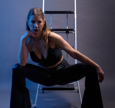 Woman in black leather outfit posing in front of a white step ladder with dramatic blue studio lighting.