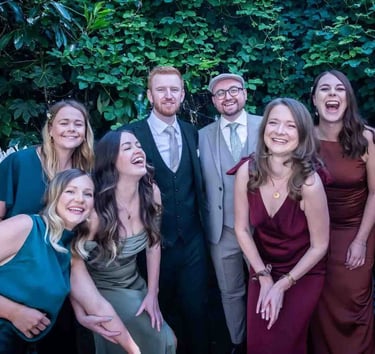 A group of happy wedding guests in formal attire laughing together in a garden setting.