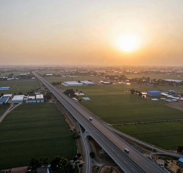 Wide panoramic aerial view of the Yamuna Expressway at sunset, showing clean multi-lane highway cutting through vast agricultural plots planned for industrial development, warm golden sunlight.