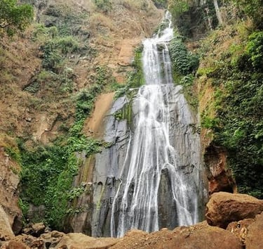 A scenic multi-tier tropical waterfall cascading down a steep rocky cliff surrounded by lush green vegetation.