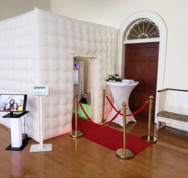 White inflatable photo booth with a red carpet and gold stanchions at a wedding reception.