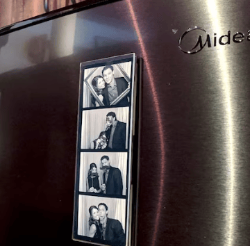 A personalized photo booth magnetic strip of a couple displayed on a black Midea refrigerator door.