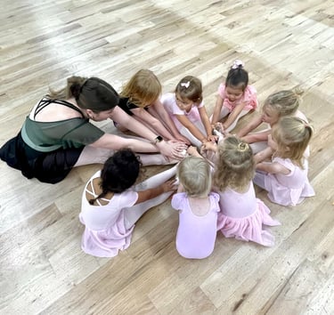 A ballet teacher and young students in pink leotards sit in a circle on a wood studio floor.