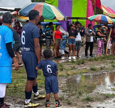 A soccer player and child in matching jerseys stand near a muddy field at a crowded community festival.