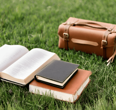 A satchel, two closed books, and an open one lay in a field of green grass