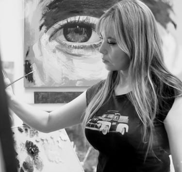 A blonde female artist paints a detailed eye portrait on canvas in a black and white studio setting.
