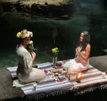 Two women seated on colorful woven blankets performing a candle-lit ritual inside a cenote in Tulum,