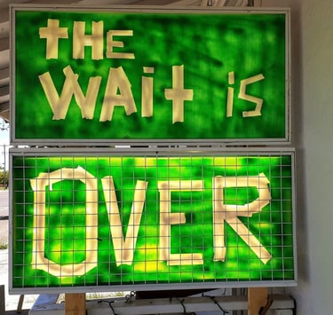 Illuminated green and yellow sign reading “The Wait Is Over.”