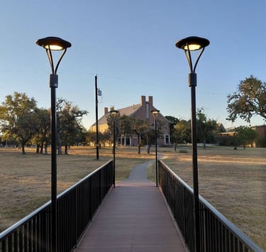 Footbridge lit by lampposts at dawn, leading toward a building in the distance.