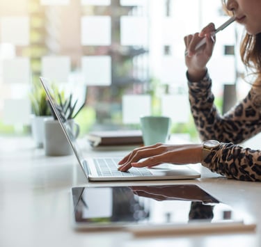 Professional woman holding a pen near her mouth, working on a laptop on a desk with a digital tablet, plant, and coffee cup.