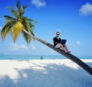 A digital nomad working on a laptop while sitting on a leaning palm tree at a tropical beach.