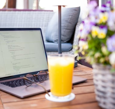 A MacBook Pro laptop displaying code on a patio table next to orange juice and flowers.
